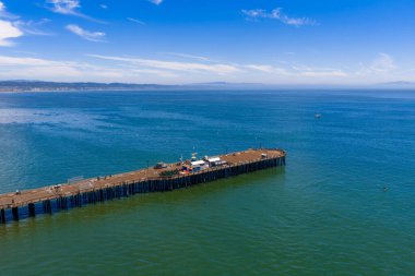 Aerial view of Capitola Pier in California, surrounded by calm blue green waters. Small boats and a distant coastline appear under a clear sky.