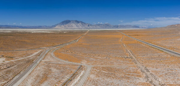 Expansive desert landscape with dirt roads, rail tracks, white salt flats, and a mountain range under a clear blue sky in Nevada.