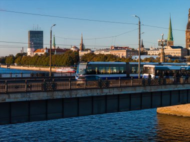 Riga, Letonya, yukarıdan bir köprüyle nehri geçerken görüldü, hareket halindeki bir tramvay ve araçlar, tarihi kuleler, ve akşam ışığında modern bir gökdelen..