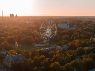 Riga 'nın güneşli bir akşamda parktaki dönme dolap, yoğun yeşillik, modern yüksek binalar ve bir iletişim kulesi görüntüsü..