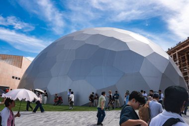 Dünya Fuarı 2025 'te mavi gökyüzünün altında altıgen ve beşgen panelleri olan büyük metalik bir jeodezik kubbe. Ziyaretçiler yakınlarda yürüyor, bazıları şemsiyeli..