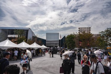 İnsanlar Osaka 'daki 2025 Dünya Fuarı' nda beyaz çadırların altında toplanıyor. Arka planda yansıtıcı cam yapı da dahil olmak üzere modern binalar yer alıyor..