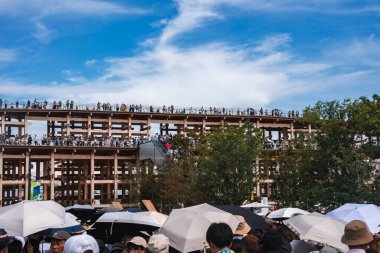 Büyük bir kalabalık mavi bir gökyüzünün altında çok katlı ahşap bir köşkün yanında toplanıyor. Katılımcılar, yeşilliklerle çevrili şemsiyelerle üst katlarda yürüyorlar..