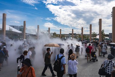 Ziyaretçiler, Japonya 'nın Osaka kentindeki 2025 Dünya Fuarı' nda, arka planda modern yapılar ve yeşilliklerle uzun tahta sütunlar ve yapay sis arasında yürürler..