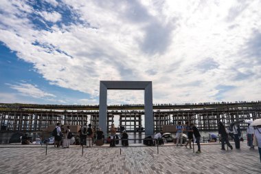 Japonya 'nın Osaka şehrinde 2025 Dünya Fuarı' nda büyük bir dikdörtgen çerçeve ve ahşap kafes geçidi. Ziyaretçiler bulutlu bir gökyüzünün altında yürür ve toplanır.