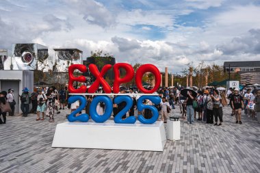 Ziyaretçiler, modern mimari ve Osaka 'da parçalı bulutlu bir gökyüzü ile çevrili beyaz bir platformda büyük kırmızı ve mavi bir EXPO 2025 tabelasının etrafında toplanıyorlar..