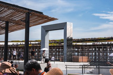 2025 Dünya Fuarı 'nda büyük metalik dikdörtgen bir kemer mavi gökyüzüne karşı duruyor. Ziyaretçiler gölgeli alanlar ve arka planda yeşil alanlar altında yürüyor..