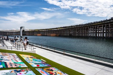 Modern bir ahşap yapı Osaka 'daki Dünya Fuarı 2025' te bir su kütlesini kapsar. Kalabalık üst katta, çimenli bir alanda renkli sanat eserleriyle yürüyor..