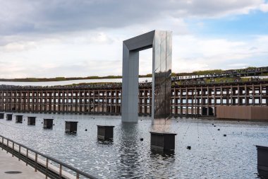 Bir su özelliğinden yansıtıcı metalik kemer yükselir. Yapı gibi ahşap bir ızgara ve bulutlu bir gökyüzünün altında kalabalıklar vardır..