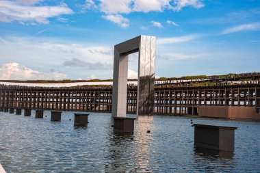 Büyük metalik dikdörtgen bir kemer, ahşap kafes yapıları, yeşil bir çatı geçidi ve ziyaretçiler ile çevrili yansıtıcı bir su gövdesi üzerinde durur..