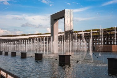 Yansıtıcı dikdörtgen kemerli bir kemer, Japonya 'nın Osaka kentindeki Dünya Fuarı 2025' te, açık mavi gökyüzünün altında senkronize fıskiyeleri olan bir havuzda duruyor..