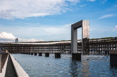 Yansıtıcı büyük bir dikdörtgen kemer, yeşil ve uzak bir bina ile kıvrımlı bir pavyonun yanında, çeşme ile bir havuz üzerinde durur..
