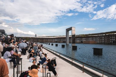 Japonya 'nın Osaka kentindeki bir rıhtımda büyük bir kalabalık toplanıyor. Metalik bir yapı, yüksek bir yürüyüş yolu ve bulutlu bir gökyüzünün altında deniz kenarında oturan ziyaretçiler yer alıyor..
