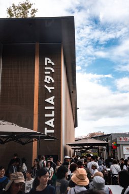 Ziyaretçiler, Japonya 'nın Osaka kentindeki 2025 Dünya Fuarı' nda İtalya Pavyonu 'nu geziyorlar. Modern yapıda parçalı bulutlu bir gökyüzü altında dikey bir 'ITALIA' işareti yer almaktadır..