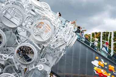 Birbirine bağlı bisikletler, aynalı yüzeyleri olan beyaz bir heykel oluşturur ve izleyicileri yansıtır. Arka planda cam merdiven ve soyut grafikler görülüyor.