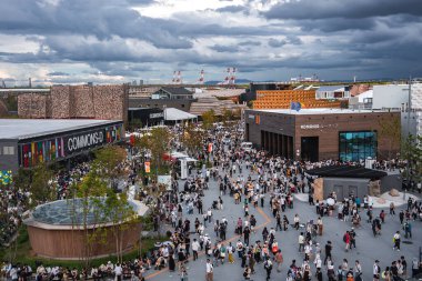 Ziyaretçiler, modern pavyonlar, bayraklar, renkli işaretler ve bulutlu bir arkaplan ile çevrili Osaka, Japonya 'daki 2025 Dünya Fuarı' nda bir plazada toplandılar..