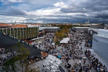 Ziyaretçiler modern bir meydanda toplandılar. Çadırlar, yeşil bir duvar cephesi ve endüstriyel ufuk çizgisi ve bulutlu gökyüzüne karşı yansıtıcı bir geometrik heykel..