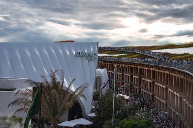 Avustralya Pavyonu 'nda ziyaretçiler, yeşil çatı, bayraklar ve Osaka' da bulutlu bir gökyüzü altında kalabalıklar ile kıvrımlı beyaz bir cephe yer alıyor..