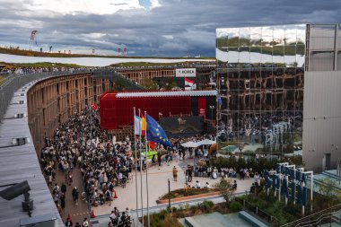 Japonya 'nın Osaka kentindeki 2025 Dünya Fuarı' nda büyük bir kalabalık toplanıyor, modern mimari, ulusal bayraklar, manzaralı yeşillikler ve bulutlu bir gökyüzü ile çevrili..