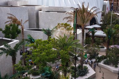 Palmiye ağaçları ve tropikal bitkilerle çevrili modern beyaz yapılar, ziyaretçiler yürüyor ve yeşil bir bayrak sergileniyor..