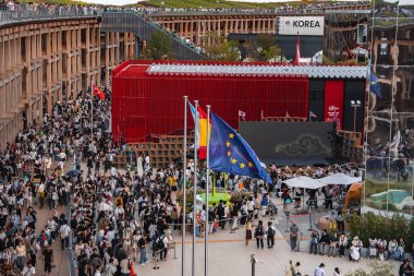 Ziyaretçiler, Japonya 'nın Osaka kentindeki 2025 Dünya Fuarı' nda kırmızı bir pavyonun yakınında toplandılar. Canlı sahnede, AB bayrağı ve Kore işareti de dahil olmak üzere bayraklar görülüyor.