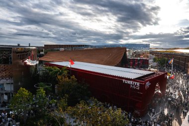 Osaka 'daki Dünya Fuarı 2025' teki Türkiye pavyonunda ziyaretçiler ve küresel unsurlarla çevrili cesur bir kırmızı dış görünüş, Türk bayrağı ve modern tasarım yer alıyor..