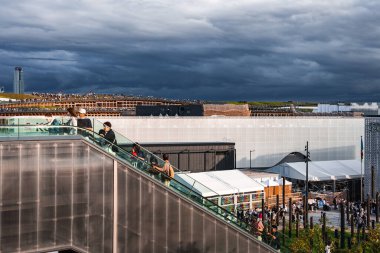 Osaka 'da 2025 Dünya Fuarı' ndan bir sahne yürüyen merdiven, beyaz pavyonlar, yeşil çatılı bir bina, uluslararası bayraklar ve sergilerle gelen ziyaretçiler..