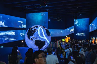 Kıvrımlı çizgileri olan parlak bir küre, dijital ekranlarla çevrili loş bir serginin ve meşgul ziyaretçi kalabalığının merkezidir..