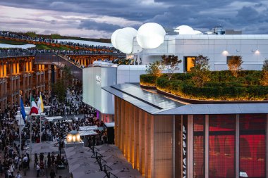 2025 Dünya Fuarı 'nda bir çatı bahçesi ve parlayan küreler modern bir yapıyı ön plana çıkarıyor. Kalabalıklar, bayraklar ve bulutlu bir akşam gökyüzü sahneyi tamamlıyor..