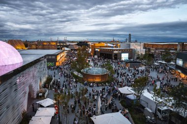 Japonya 'nın Osaka şehrindeki 2025 Dünya Fuarı' nda ziyaretçiler, modern mimari, ağaçlar, köşkler ve arka planda bulutlu bir gökyüzü ile çevrili açık bir meydanı dolduruyor..