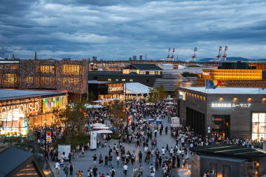 Ziyaretçiler Japonya 'nın Osaka kentinde 2025 Dünya Fuarı' nı gezerken, sağda Romanya pavyonu, solda Avam Kamarası D ve arka planda bulutlu bir şehir manzarası bulunuyor..