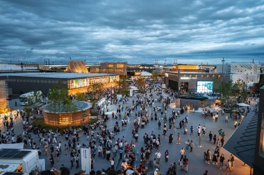 Ziyaretçiler, modern binalar, canlı tabelalar, dairesel bir pavyon ve bulutlu bir rıhtım zemininin yer aldığı Osaka, Japonya 'daki 2025 Dünya Fuarı' nı keşfediyorlar..