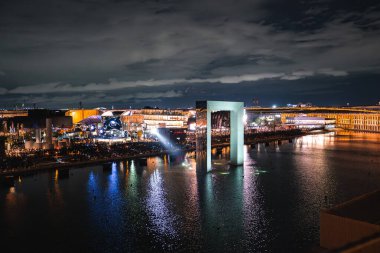 Aydınlatılmış kemer yapısı Osaka 'daki Dünya Fuarı 2025' teki suya yansıyor. Modern pavyonlar, kalabalık ve parçalı bulutlu gökyüzü canlı bir sahne yaratır..