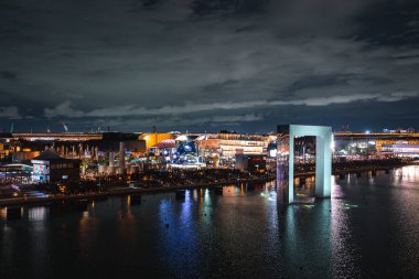 Japonya 'nın Osaka kentinde 2025 Dünya Fuarı' nın gece görüşü, su, modern mimari ve bulutlu bir gökyüzü çizgisi üzerinde geniş aydınlatılmış bir kemer içerir..