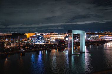 Osaka 'daki Dünya Fuarı 2025' te canlı bir gece sahnesi. Suyun, modern mimarinin ve hareketli bir kalabalığın üzerinde dikdörtgen bir kemer var..