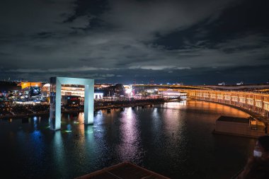 Japonya, Osaka 'da 2025 Dünya Fuarı' nın canlı bir gece sahnesinde su, modern binalar ve hareketli kalabalıklar üzerinde yanan dikdörtgen bir kemer yer alıyor..