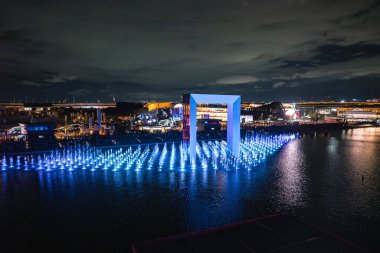 2025 Dünya Fuarı 'nda mavi fıskiyeli parlayan bir havuzun üzerinde modern pavyonlar ve renkli ışıklarla çevrili büyük, aydınlık bir kemer duruyor..