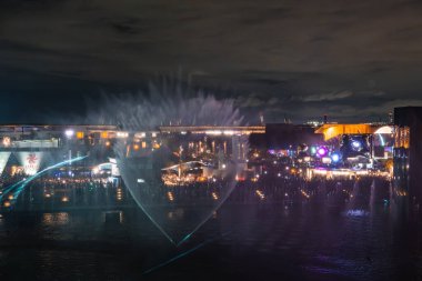 Japonya 'nın Osaka kentinde 2025 Dünya Fuarı' nda, renkli ışıklar, modern pavyonlar ve hareketli kalabalıklarla aydınlatılmış büyük bir su çeşmesinin gece manzarası..