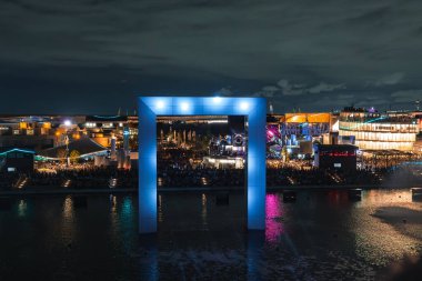 Osaka 'daki 2025 Dünya Fuarı' nda yansıtıcı suyun üzerinde büyük mavi bir kemer duruyor. Etrafı modern pavyonlar, renkli ışıklar ve kalabalıklarla çevrili..