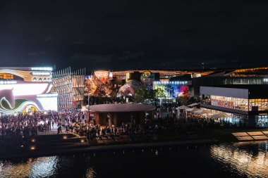 Japonya 'nın Osaka şehrinde 2025 Dünya Fuarı' nda, parlak fütürist pavyonlar, dairesel bir plaza ve parıldayan su yansımalarının yer aldığı canlı bir gece sahnesi..