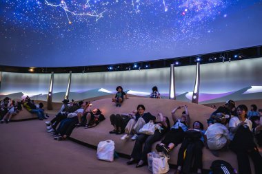 İnsanlar, modern sergi alanında, kıvrımlı koltuklarda, kubbe bir ekranla, takımyıldızları göstererek rahatlar. Oda parlak, fütüristik bir tasarıma sahip..