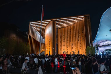 Dünya Fuarı 2025, Osaka 'da altın ışıklandırma ve hat panelleri olan eğri bir pavyon. Kırmızı bayrak ve canlı bir kalabalık coşkulu sahneyi canlandırıyor..