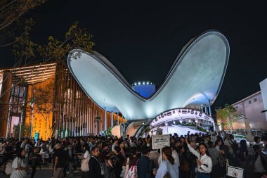 Aydınlatılmış kelebek şekilli bina, Osaka 'daki Dünya Fuarı 2025' te eğri kanatlı. Kalabalıklar, ağaçlar ve ahşap bir yapı canlandırıyor sahneyi.