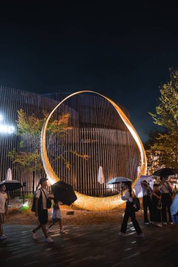 Japonya 'nın Osaka şehrinde 2025 Dünya Fuarı' nda, modern mimari, ağaçlar ve ahşap bir yolda ziyaretçiler tarafından çevrelenmiş parlak dairesel bir heykelin gece görüntüsü..