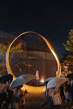 Dünya Fuarı 2025, Osaka, Japonya 'daki parıldayan altın halka yapısının gece manzarası, modern mimari, seyrek ağaçlar ve yağmurlu bir kalabalık..