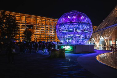 Dünya Fuarı 2025 'te mor ve mavi ışıklarla aydınlatılmış jeodezik küre, modern pavyonlar, ziyaretçiler ve parlayan su özelliği ile çevrili..