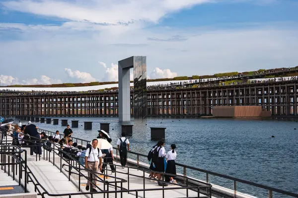 Japonya 'nın Osaka kentindeki parçalı bulutlu bir gökyüzünün altında yürüyen ve oturan ziyaretçilerle, ahşap kirişlerle çerçevelenmiş, sudan metalik bir yapı yükselir..