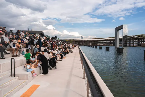 İnsanlar, Japonya 'nın Osaka kentindeki parçalı bulutlu bir gökyüzünün altında yer alan, metalik bir yapısı ve yükseltilmiş yürüyüş yolu olan bir rıhtımın yanındaki katlı basamaklarda toplanıyorlar..