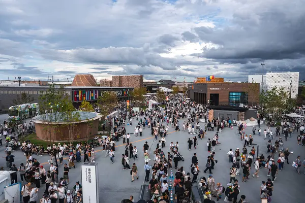 Ziyaretçiler, modern pavyonlar, dairesel ahşap bir bina ve ROBINSON etiketli bir tuğla bina ile çevrili, Dünya Fuarı 2025 'te açık bir plazada toplandılar..