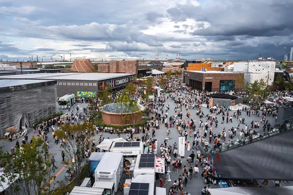 Ziyaretçiler modern pavyonlar, yeşillikler, yemek kamyonları ve çatı bahçesi olan bir meydanda toplanıyorlar. Vinçler ve depolar bulutlu bir gökyüzünün altında beliriyor.